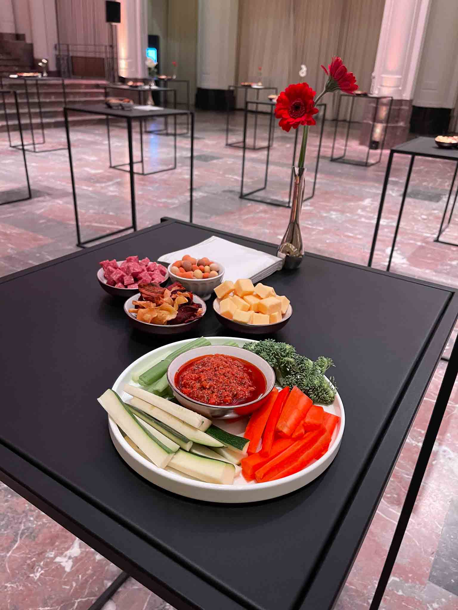 Een zwarte tafel met daarop een bord met celder, wortel, brocolli en een dipsausje in het midden. Daarachter meerdere kleine kommetjes met borrelhapjes waaronder nootjes, kaas, salami en chips.